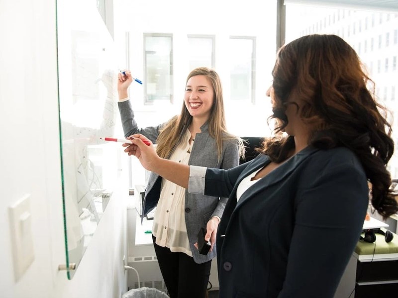 Two women standing at a whiteboard, writing something and looking happy.