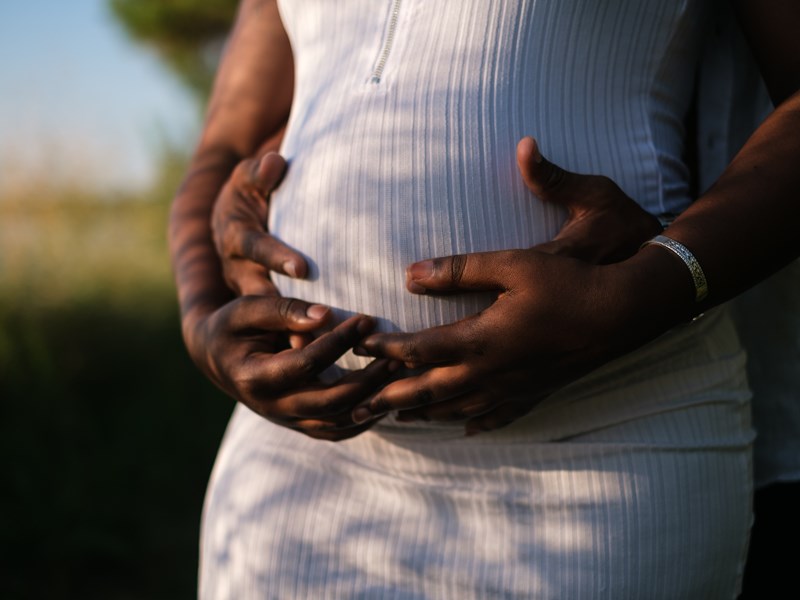 A pregnant lady in a white dress with her partner, both holding onto her baby bump.